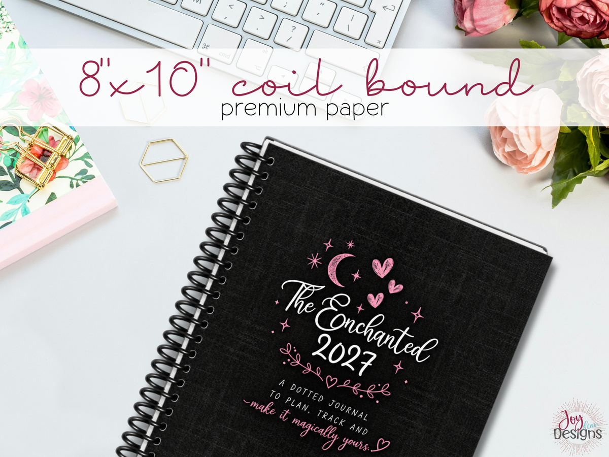 The Enchanted 2027 physical dotted journal shown with an 8 x 10 inch softcover, black coil binding and premium paper. The black linen-look cover features white lettering, pink hearts, crescent moon and star illustrations. This coil-bound planner opens flat for comfortable writing, tracking, colouring and journaling. A ready-to-use dated 2027 journal with over 350 pages of calendars, weekly spreads, trackers and creative layouts.