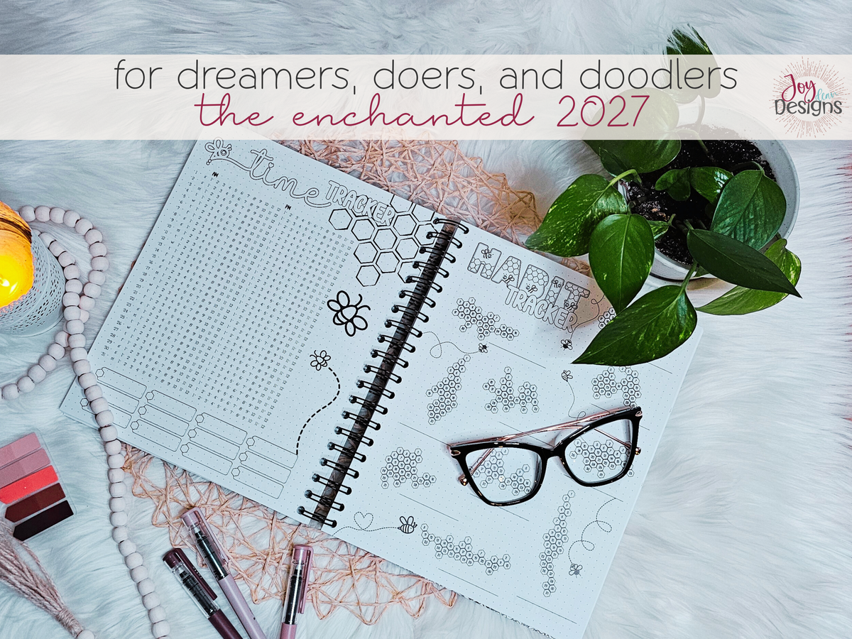 Open coil-bound dotted journal displaying a two-page time tracker and habit tracker spread with illustrated bees and honeycomb designs. The black wire binding allows the journal to lie open for writing and colouring. Photographed with pens, eyeglasses, greenery and a candle on a soft white background. An example of the detailed pre-designed tracking pages included in The Enchanted 2027 creative planner by Joy Dean Designs.