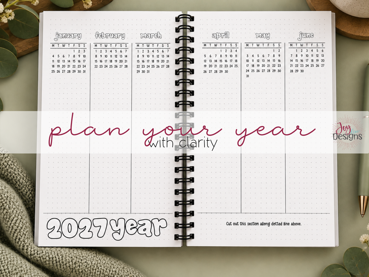 Open coil bound The Vital 2027 planner showing the year-at-a-glance Dutch door calendar pages with January through June visible. The black and white dotted journal features miniature monthly calendars and spacious planning columns for organizing the year. Displayed with the words Plan Your Year With Clarity on a cozy sage green desktop with flowers, knitted fabric and stationery.