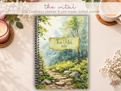 The Vital 2027 wellness planner and pre-made dotted journal by Joy Dean Designs, featuring a beautiful watercolor forest pathway cover with green trees, flowers and a black coil binding. Displayed on a cozy cream desk with coffee, candles and flowers. This dated 2027 planner combines weekly planning, self-care, health and wellness tracking, habit trackers, mood trackers and creative coloring pages in one spiral bound journal.
