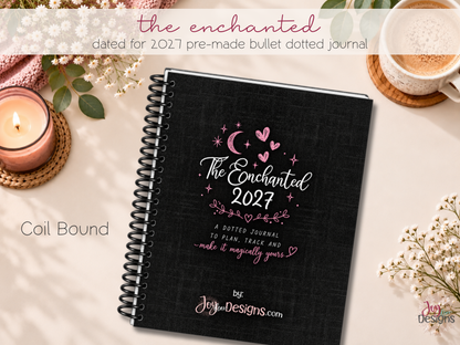 The Enchanted 2027 pre-made dotted journal, an 8 x 10 inch softcover planner with black coil binding and a black linen-look cover decorated with pink hearts, a crescent moon and stars. This dated 2027 journal is designed for creative planning, organization, tracking habits and everyday journaling. A ready-to-use planner with pre-designed pages, monthly themes and space for personal notes, doodles and colouring. Displayed on a desk with flowers and stationery.